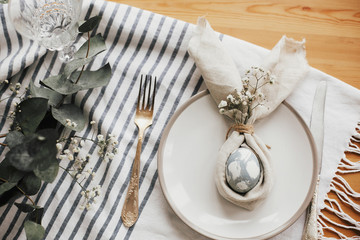 Easter table decorations. Stylish Easter brunch table setting with egg in easter bunny napkin. Modern natural dyed blue egg on napkin with bunny ears, flowers on plate and vintage cutlery.