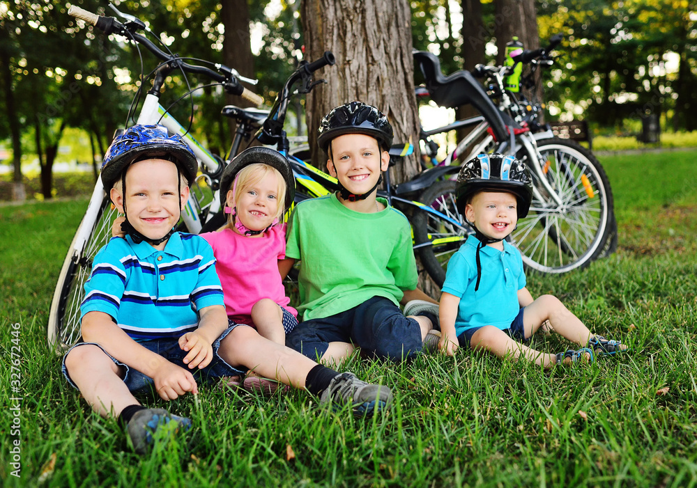 Fototapeta premium a group of small preschool children in Bicycle safety helmets smile sitting on the fresh green grass in the Park against the background of bicycles and trees.