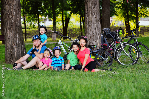 a large family-mom, dad and a group of small children sitting on the grass in Bicycle gear and helmets smiling against the background of bicycles, Park and green grass.