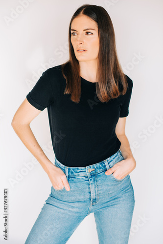 Indoor portrait of beautiful young woman, wearing black t-shirt and high waist jeans, posing on white background, holding hands in pockets