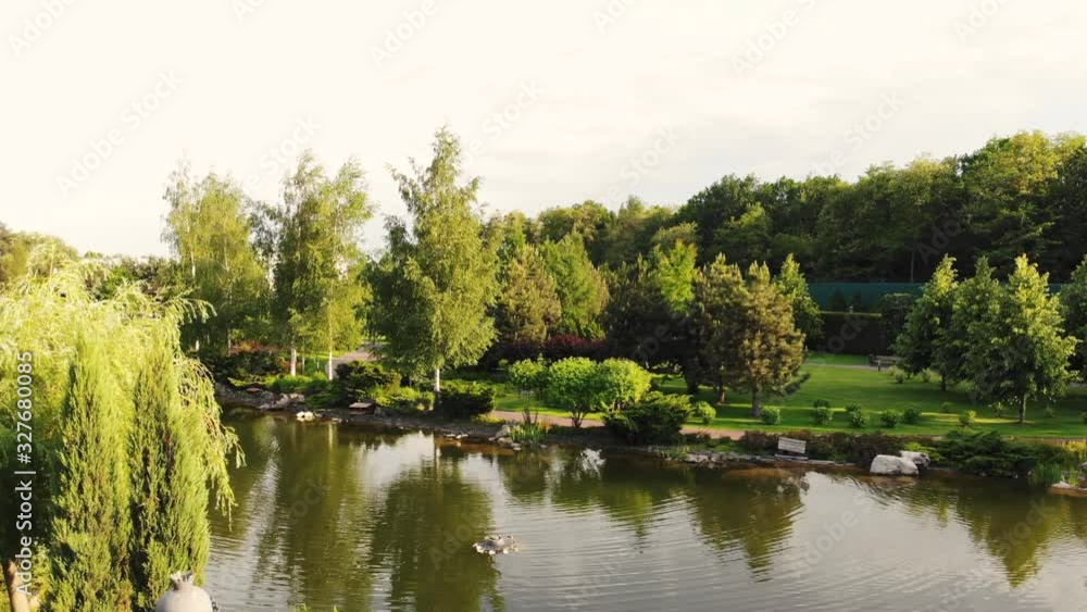 Backward fly over park pond. Landscape of well-groomed city park and reflective water surface. Backward fly over park pond. Landscape of well-groomed city park and reflective water surface.