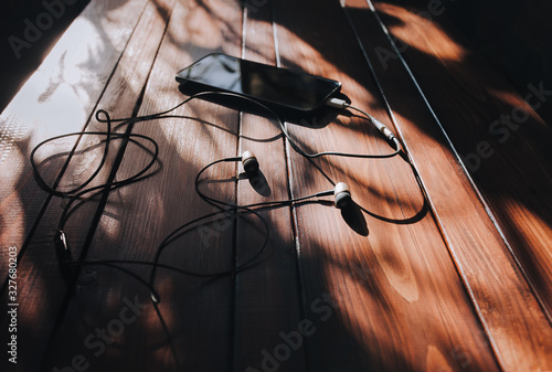Black smartphone and dark headphone lies on a brown wooden background mahogany with spots of sunlight. Close up, copy space. Morning music concept.