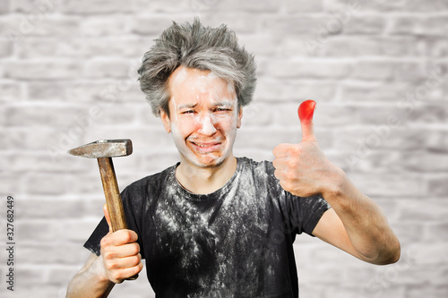 Dirty young builder guy fail is hold a hammer, hit the finger on brick wall background at home during repairs photo