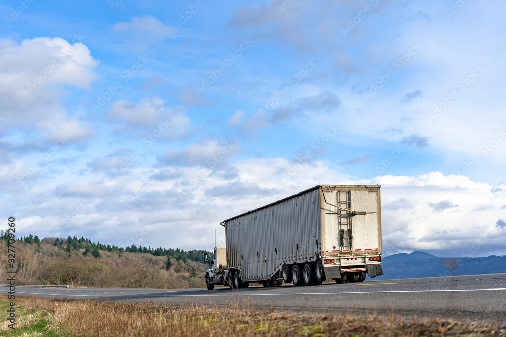 Day cab big rig semi truck transporting cargo in covered corrugated bulk semi trailer driving on the road with frees on the hill Day cab big rig semi truck transporting cargo in covered corrugated bulk semi trailer driving on the road with frees on the hill