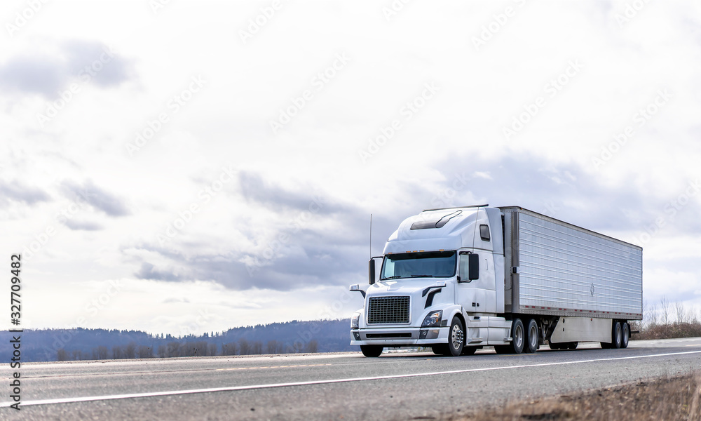 Obraz premium Industrial transportation big rig white semi truck transporting frozen cargo in reefer semi trailer running on the road with cloudy sky