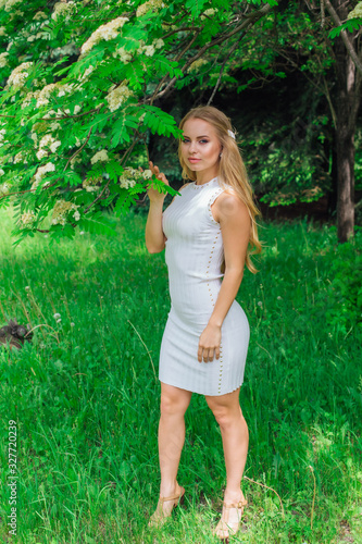 Portrait of a charming blond woman wearing beautiful white dress standing next to rowan tree with white flowers. photo