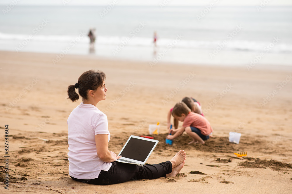 Relaxed mom teleworking at the beach as children play with sand