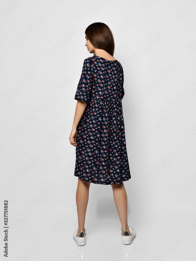 Young beautiful woman is posing backwards in new fashion long casual summer dress and white sneakers on gray Young beautiful woman is posing backwards in new fashion long casual summer dress and white sneakers on gray