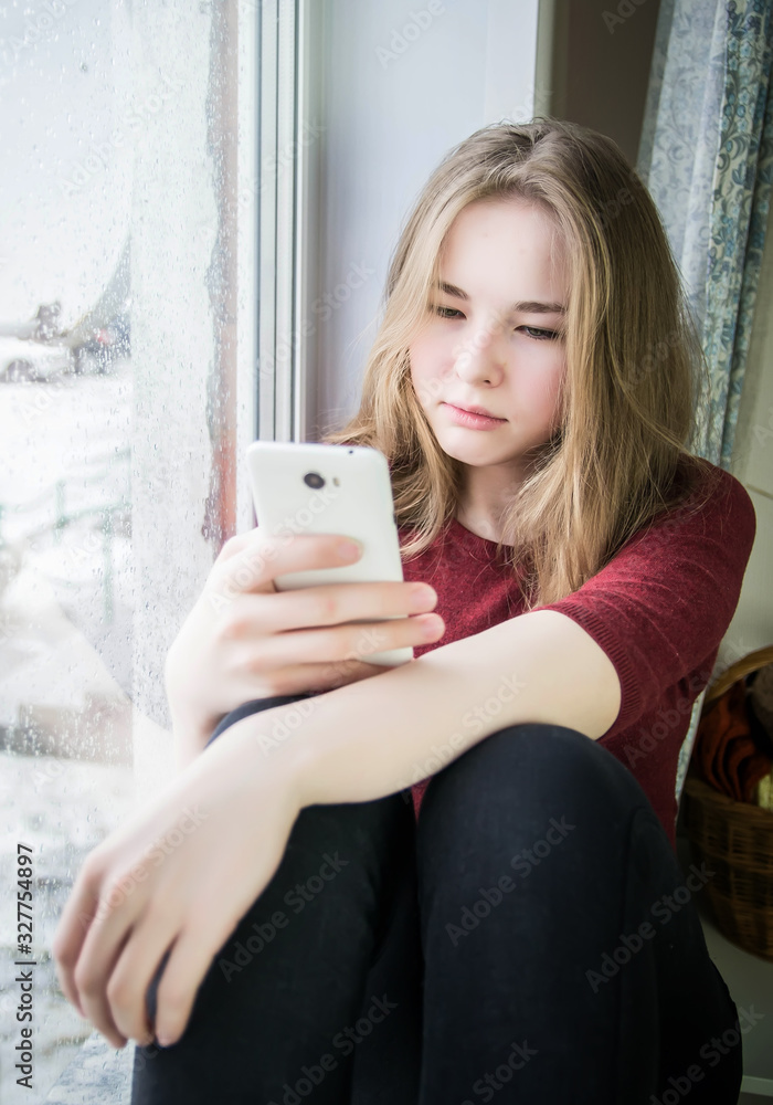 Overweight teenager girl with long blond hair is sitting by the window on the windowsill with a phone in her hand. The concept of teenage addiction to a mobile phone, the Internet, depression Overweight teenager girl with long blond hair is sitting by the window on the windowsill with a phone in her hand. The concept of teenage addiction to a mobile phone, the Internet, depression