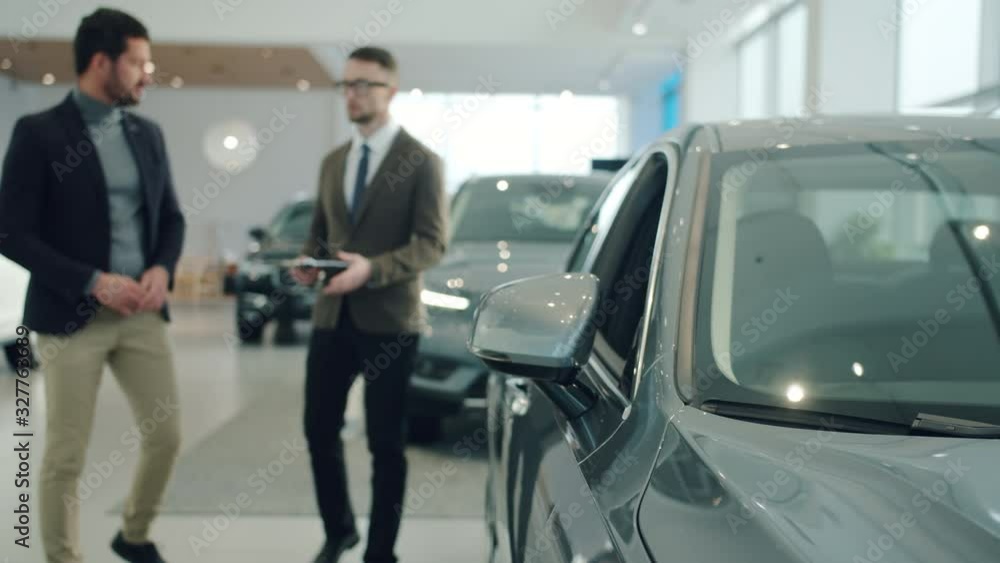 Male buyer businessman is choosing brand new car in showroom discussing deal with manager walking and talking indoors. People and business concept. Focus on vehicle mirror Male buyer businessman is choosing brand new car in showroom discussing deal with manager walking and talking indoors. People and business concept. Focus on vehicle mirror