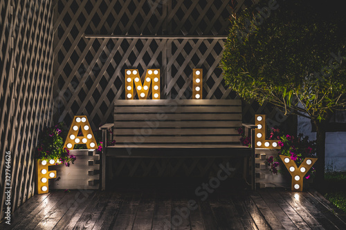 The word FAMILY is decorated with glowing letters. There is a bench between the letters . There is a wooden fence at the back and a green tree at the side. Night