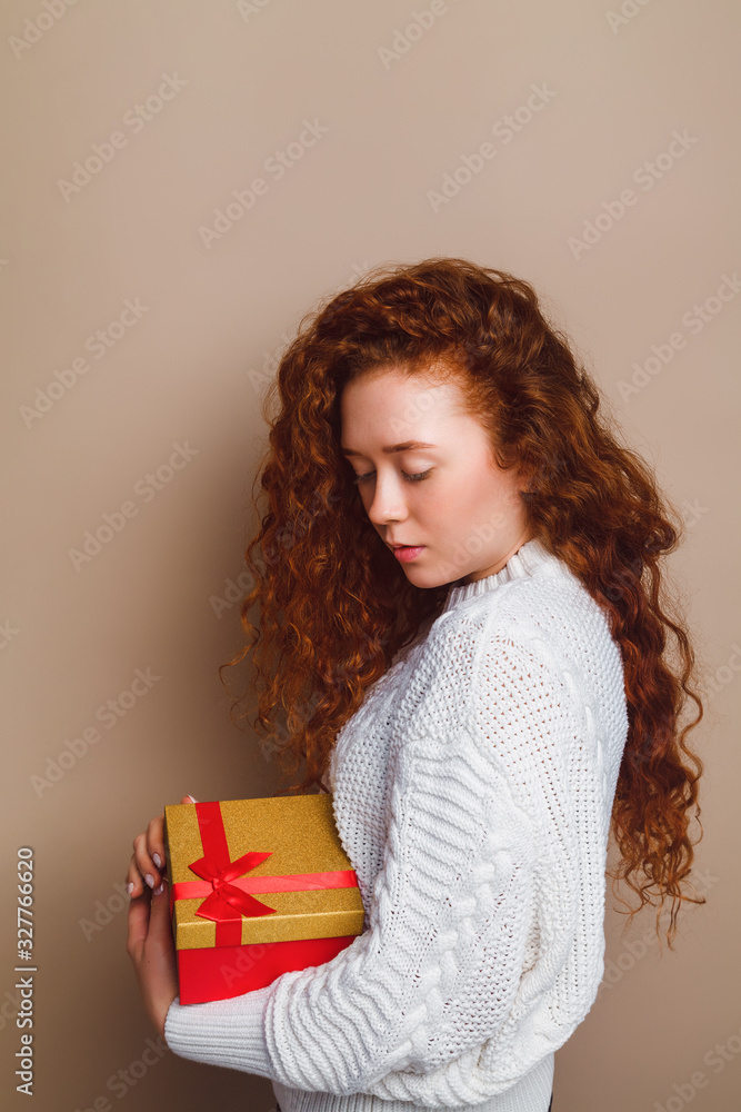 red-haired girl in a white sweater holds a red-golden gift box in her hands. Shot in a studio on a beige background. Copy space for your text, vertical red-haired girl in a white sweater holds a red-golden gift box in her hands. Shot in a studio on a beige background. Copy space for your text, vertical