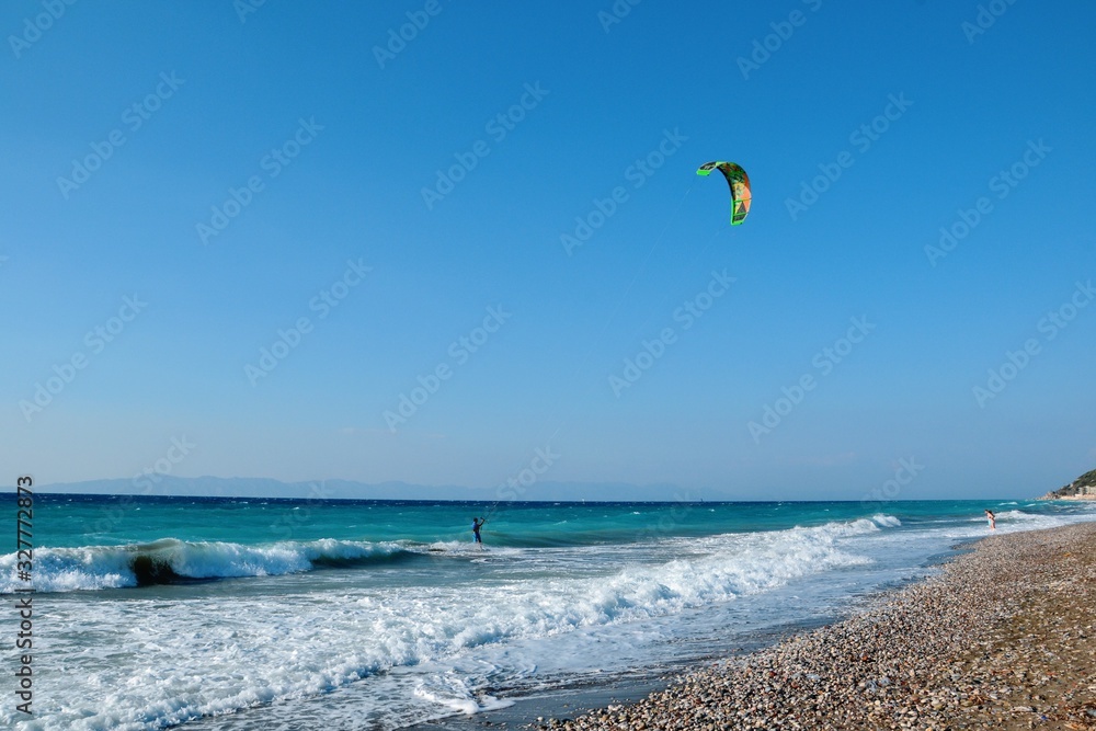 Ixia, Rhodes, Greece. A kiteboarder harnesses the power of the wind with a large controllable power kite to be propelled across the sea water. Pebble beach in Paralia/Ixia Ixia, Rhodes, Greece. A kiteboarder harnesses the power of the wind with a large controllable power kite to be propelled across the sea water. Pebble beach in Paralia/Ixia