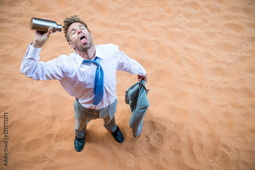 Thirsty businessman standing on red sand desert pouring an empty water bottle over his tongue