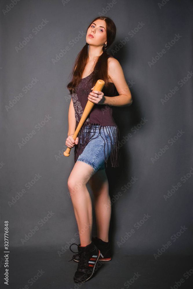 Young beautiful woman with a baseball bat in her hands. Fashionable studio photo on a gray background in the gangster style. Casual wear. Model with clean skin. Young beautiful woman with a baseball bat in her hands. Fashionable studio photo on a gray background in the gangster style. Casual wear. Model with clean skin.