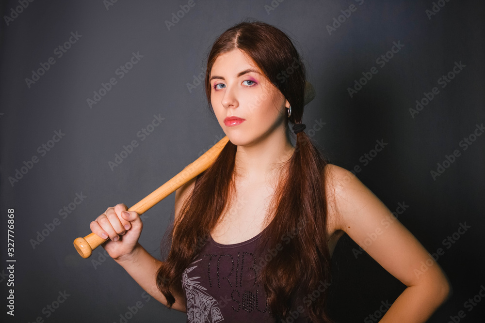 Obraz premium Young beautiful woman with a baseball bat in her hands. Fashionable studio photo on a gray background in the gangster style. Casual wear. Model with clean skin.