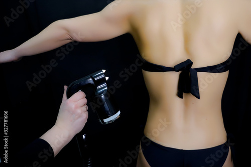 hand of beautician with airbrush-pistol for spray tan applying to young female back wearing bikini in beauty salon, Woman body paint with airbrush in professional beauty salon
