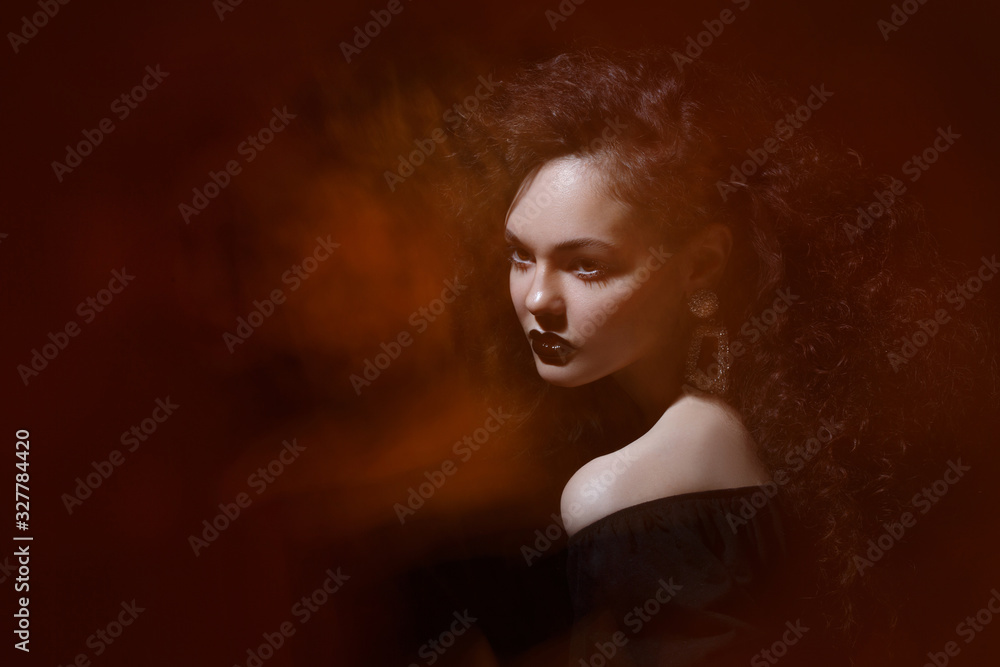 Beauty portrait of a beautiful girl with black lipstick on her lips and with curly curls on a dark background with an orange glow, mixed light.