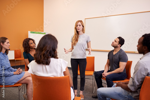 Woman Standing To Address Group Of Men And Women At Mental Health Group Therapy Meeting