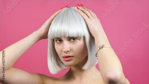 Cute young pretty woman model in blond bob wig without makeup posing in the studio on a pink background, straightens her hair, attracts attention. close-up