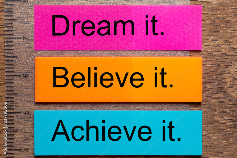 Three bookmark stickers with the words dream it, believe it, achieve it, on a dark natural wooden table
