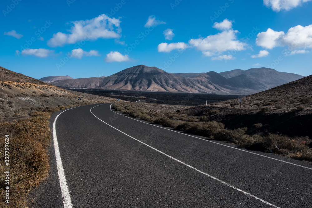 Endless road to Timanfaya National park in Lanzarote, Canary Ispands, Spain, Europe, Africa. Volcanic, black sand, harsh, tough, inhospitable, dry, sub-tropical, desert landscape. Endless road to Timanfaya National park in Lanzarote, Canary Ispands, Spain, Europe, Africa. Volcanic, black sand, harsh, tough, inhospitable, dry, sub-tropical, desert landscape.