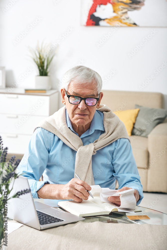 Portrait of serious modern senior man in glasses sitting at table and analyzing checks while calculating expenses Portrait of serious modern senior man in glasses sitting at table and analyzing checks while calculating expenses