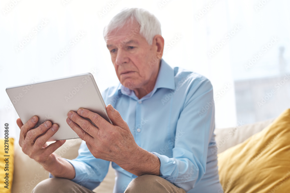 Below view of serious elderly man in shirt sitting on sofa and watching movie with interest on tablet Below view of serious elderly man in shirt sitting on sofa and watching movie with interest on tablet