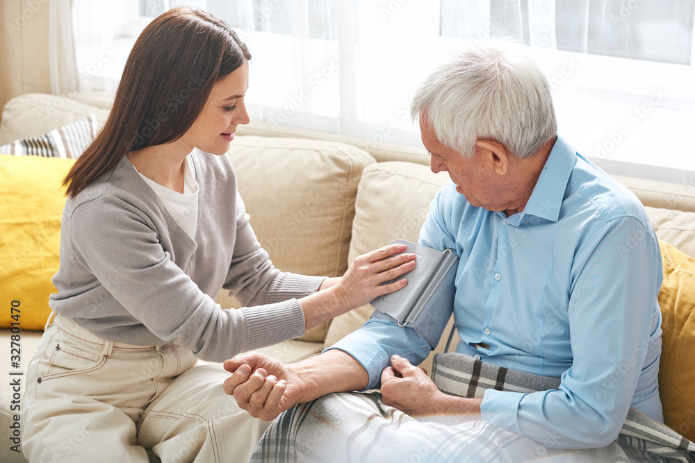 Young nurse sitting on sofa and taking pressure of elderly man in living room, she visiting patient at home Young nurse sitting on sofa and taking pressure of elderly man in living room, she visiting patient at home