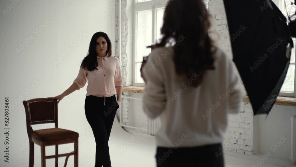 Young beautiful woman model posing with chair in photo studio for female photographer while she is shooting with digital camera. Concept of creative work in photo studio. Shooting in slow motion. Young beautiful woman model posing with chair in photo studio for female photographer while she is shooting with digital camera. Concept of creative work in photo studio. Shooting in slow motion.
