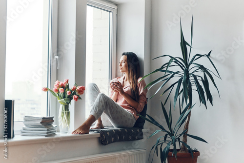 Thoughtful young woman in cozy pajamas looking through the window while resting at home photo