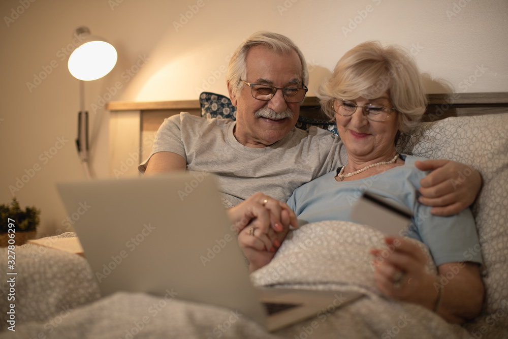 Happy senior couple buying on the Internet while using laptop and credit card in bedroom. Happy senior couple buying on the Internet while using laptop and credit card in bedroom.