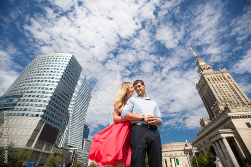 Bottom view. Woman in red dress hugs her husband on the background of the city and blue sky with clouds. Poland, Warsaw.