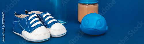 panoramic shot of baby bottle, toy, baby shoes, jar with baby food ob blue background