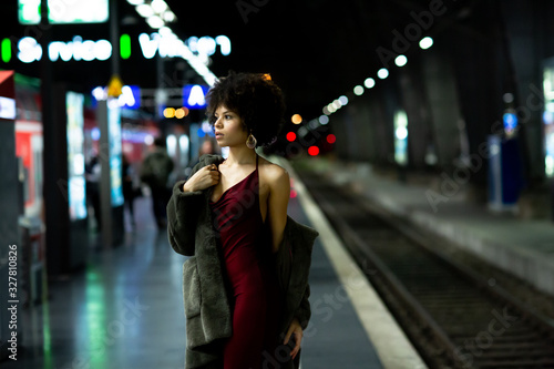 Young stylish African-American model woman in red dress and a fur coat posing on the subway platform at night. Fashion and lifestyle concept. Nightlife in the city