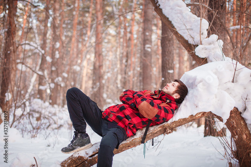 Photo of cheerful male resting, looks directly into camera, sits in snow pine winter forest, smiles happily, enjoys communication, admires landscapes