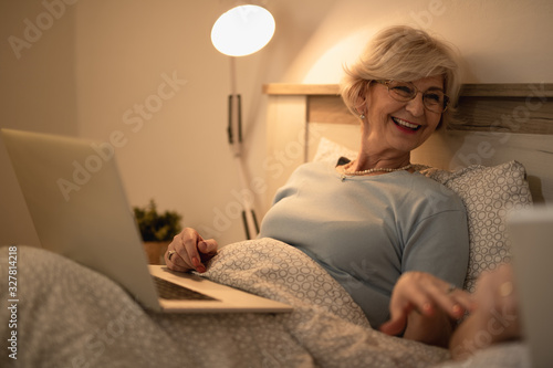 Happy mature woman talking to her husband while using laptop in bed.