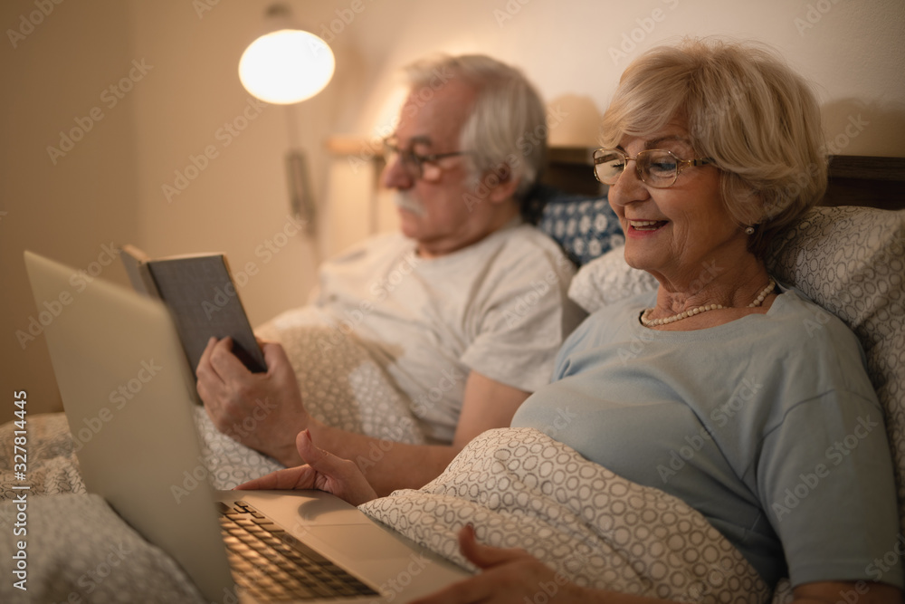 Happy senior woman using laptop while resting in bed with her husband.