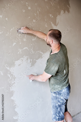 Male worker plastering wall with putty-knife, close up. Fixing wall surface and preparation for painting. Young man with a beard in a T-shirt and shorts smeared with paint