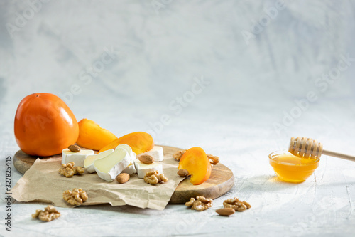 Brie and persimmon with almonds and walnuts on a wooden Board and a gray background. Honey in a glass bowl.Space for text