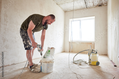 Worker mixing dry mortar with handheld mixer in bucket at repairable room. Man in smeared clothing kneads a solution against the background of a window in daylight