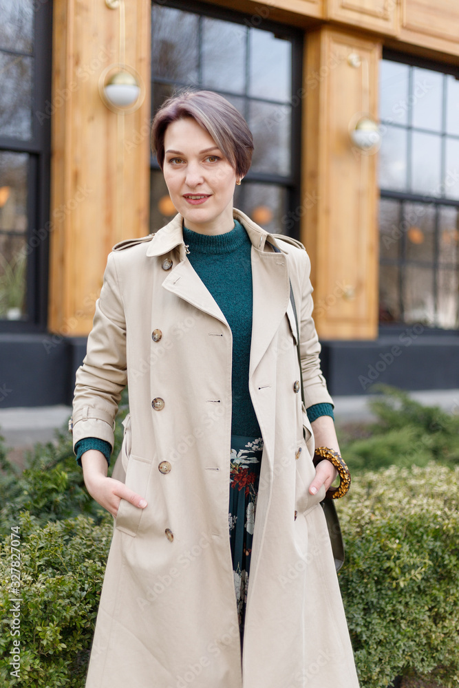 Portrait shooting of a attractive stylish short cut woman, Girl walks in the city outdoors. Stylish modern and feminine image, style. Girl in a beige cloak or coat and a green dress