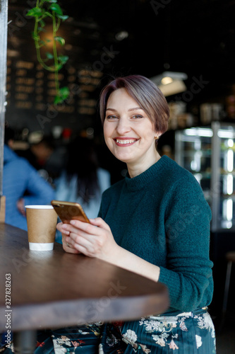 Beautiful cute girl in the cafe looking at the camera with coffee smiling. Green knitted sweater. fashionably dressed girl. girl looks at the phone. business lady