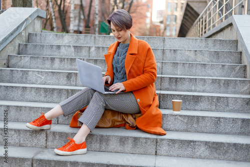 Stylish lady in bright orange coat in a city scene, using a laptop computer and working on it during a break. Outdoors technology. drinking coffee, tea, cocoa © Misha