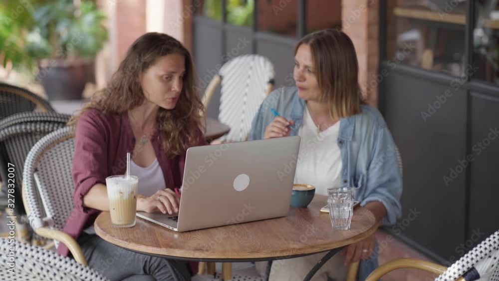 Happy female friends sitting at table in coffee shop, look at laptop screen and discuss. Girl taking notes in notebook. On table laptop and cups of coffee Happy female friends sitting at table in coffee shop, look at laptop screen and discuss. Girl taking notes in notebook. On table laptop and cups of coffee