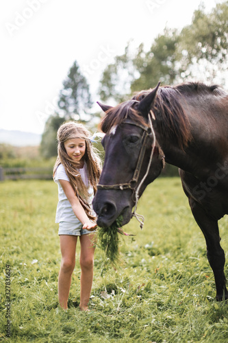 A cute girl in stylish casual clothes and hair accessories feeding her horse with green grass, in the field on a sunny summer day