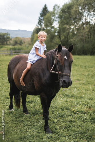 Little blond child girl in dress riding a horse. Girl on a horse walk in nature, summer field, green trees on the background. Little girl in dress galloping on her dark horse