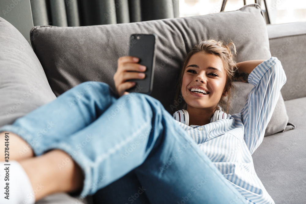 Image of smiling nice woman using cellphone while lying on sofa - obrazy, fototapety, plakaty 