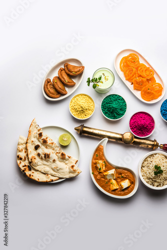 Happy Holy concept showing Indian assorted lunch food like chicken, paneer butter masala, naan, jeera rice, black chana fry, jalebi, fujiyama, thandai and Farsan with holi colours and pichkari 