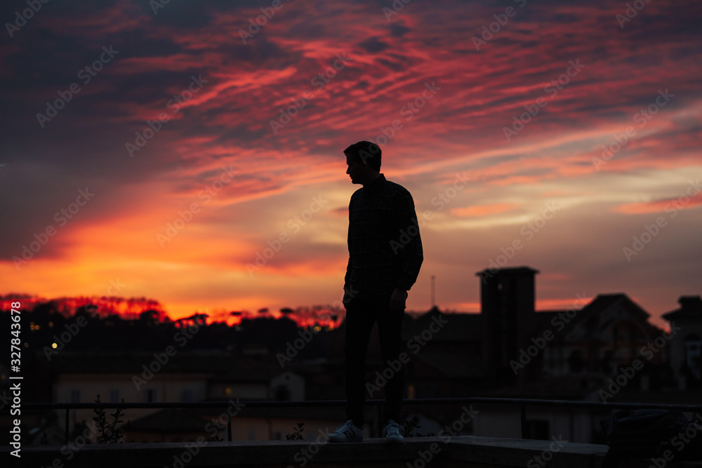 Fototapeta premium Silhouette of man standing and looking the vision over the cityscape background at sunset time. Orange, rose and blue clouds.